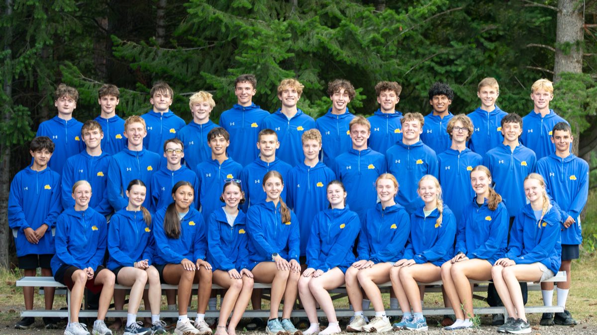 The Sheridan High School cross country team — about thirty athletes in blue and gold team jackets, standing and seated in three rows in front of pine trees.